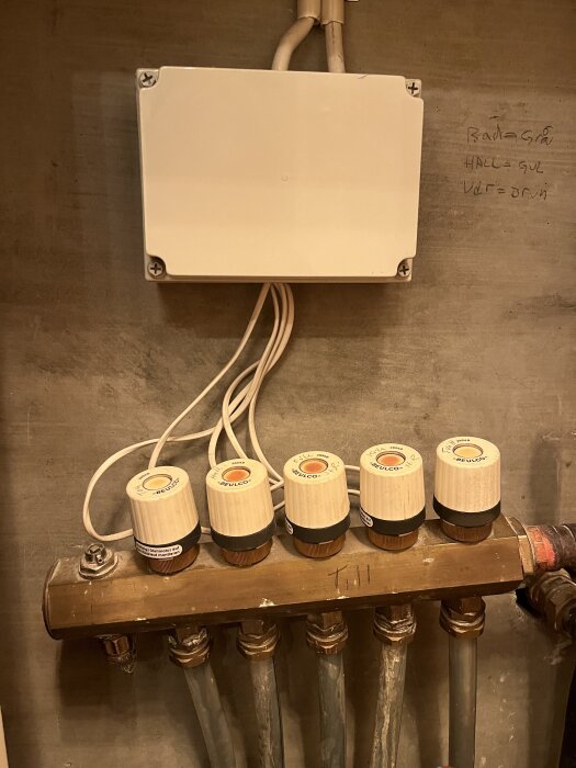Golvvärmefördelare med termostater och ställdon i teknikrum, med handskrivna etiketter för rum som bad och hall.