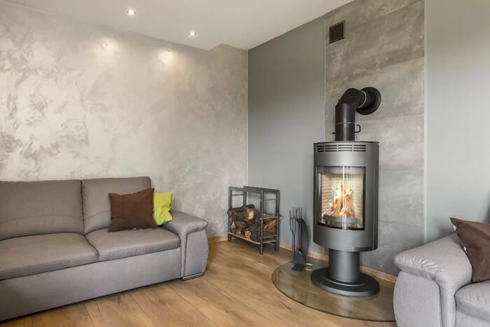 Modern kamin i hörnet av ett vardagsrum med rökrör mot murstocken och vedförvaring bredvid, omgiven av två soffor.