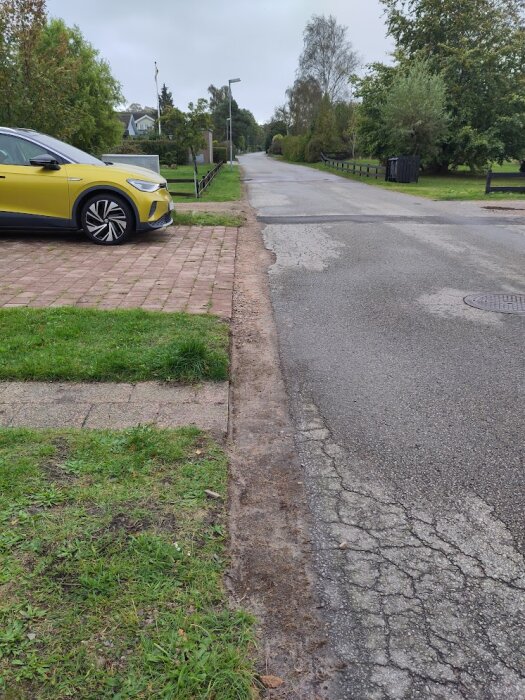 Nyasfalterad väg som möter uppfart med stenbeläggning, gul bil parkerad på uppfarten, omgiven av grönt gräs och träd i bostadsområde.