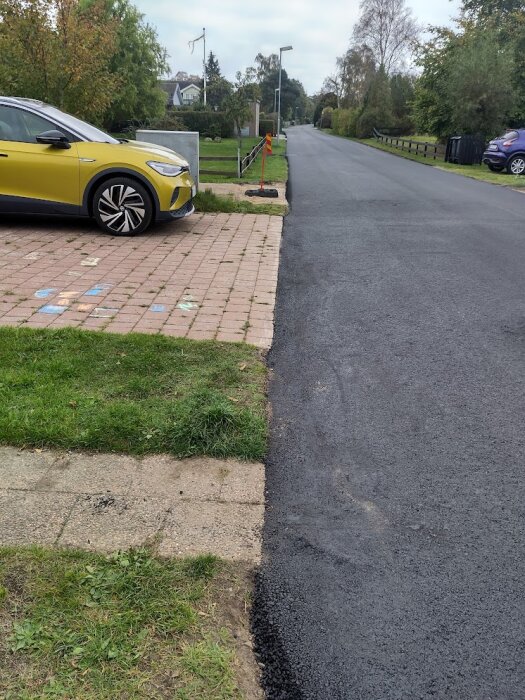 Asfalterad infart som ansluter till uppfart med stenplattor. En gul bil står parkerad på uppfarten.