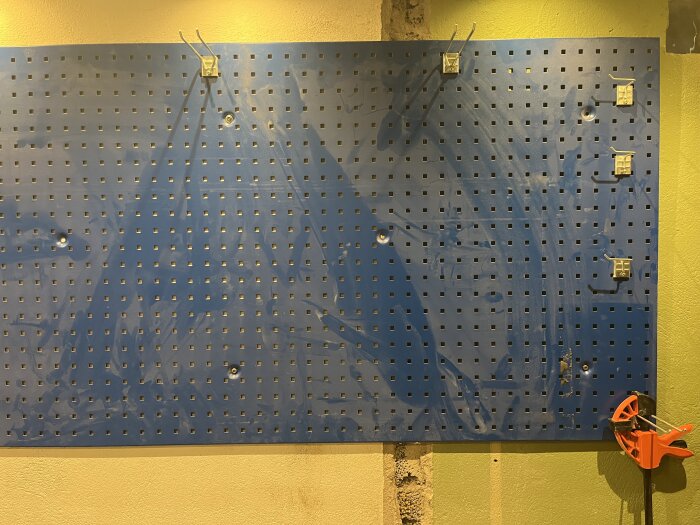 Blå verktygstavla med fyrkantiga hål och några monterade krokar, fäst på en grön vägg. En orange klämma hänger längst ner till höger.