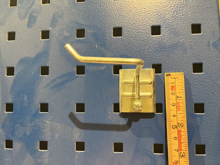 Blå verktygstavla med kvadratiska hål, monterad krok, och måttstock visar avstånd mellan hålen.