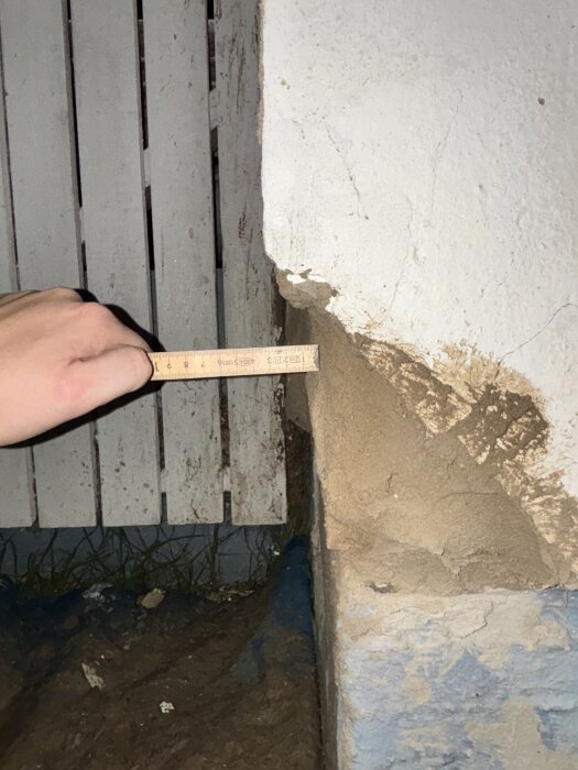 Hand mäter tjockleken på bruklagret på en slätputsad gavel, med måttstock som visar 1-4 cm, mot en vit bakgrund och träribbor.