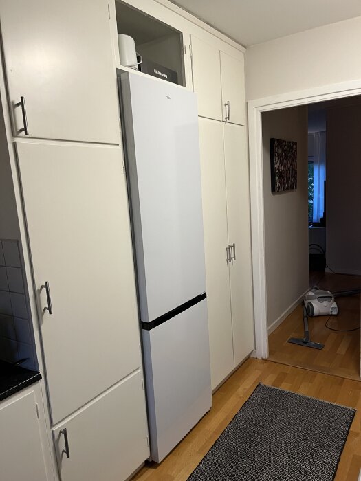 Vit kyl och frys inbyggd i vita köksskåp med träinslag på golv. En dammsugare syns i bakgrunden.
