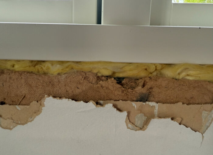 Hål i vägg runt nytt fönster i stuga, med gammalt isoleringsmaterial och pappskivor synliga.