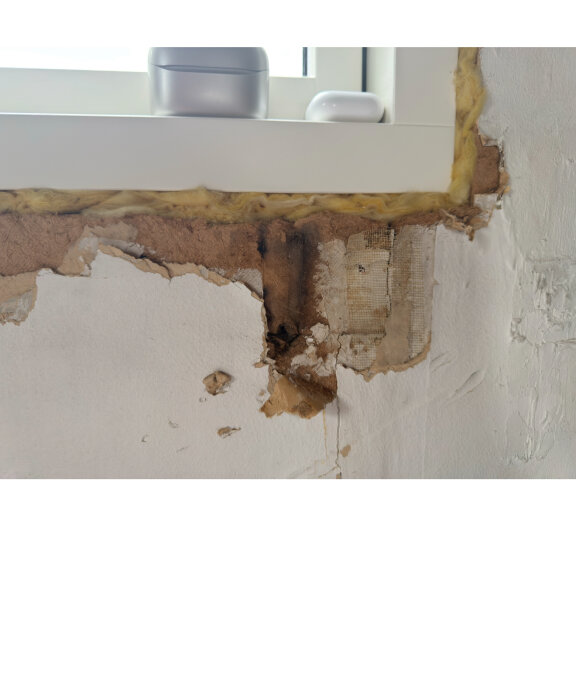 Hål i vägg vid fönsterkarm, gammalt isoleringsmaterial med papp synligt, slitage och fuktskador på väggen. Möjlig reparation diskuterad.