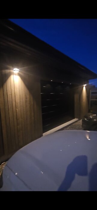 Upplyst garage exteriör på kvällen, med brädväggar och en stängd dörr; del av en bil synlig i förgrunden.