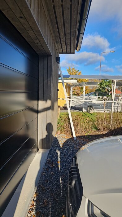 Skugga av en person mot en garagedörr, bredvid ett parkerat fordon, med ett gult hus och träd i bakgrunden under en blå himmel.