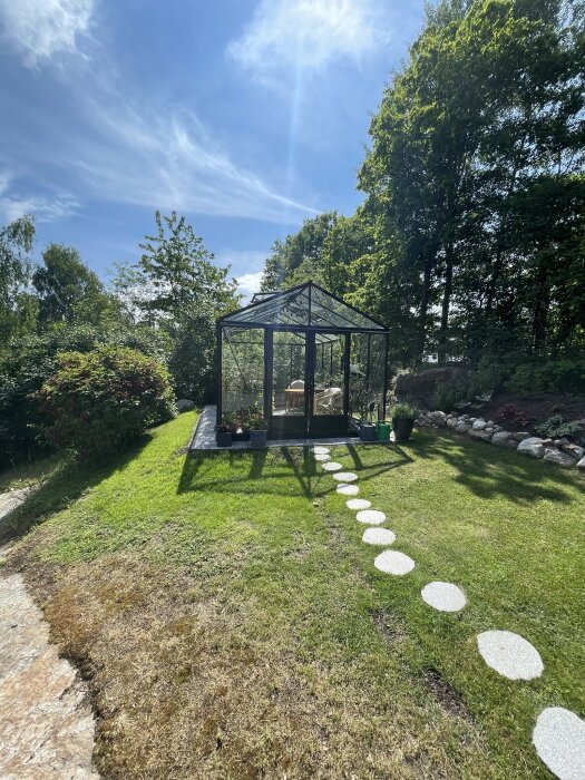 Växthus i glas beläget i en grön trädgård med en stig av runda stenar, omgiven av grönska och blå himmel i bakgrunden.