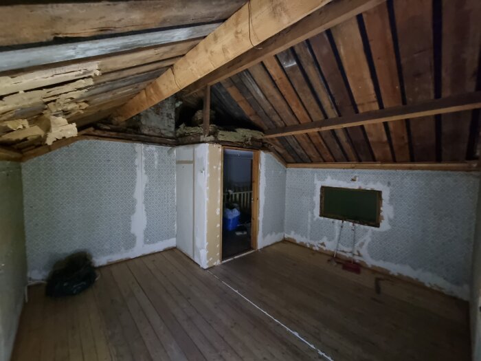 Renoveringsprojekt i 1900-talstorp; rum med synligt innertak och träpaneler, vita fläckar på väggen, liten dörr och fönster synliga i bakgrunden.