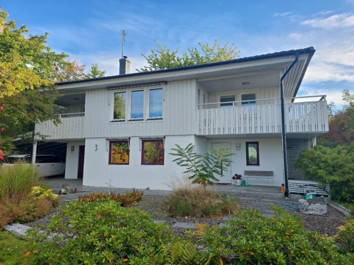 Vit tvåvåningsvilla med balkong och carport, omgiven av trädgård med grönska och grusgångar.