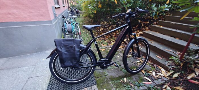 Svart cykel med en väska parkerad på en matta nära en trappa och väg.