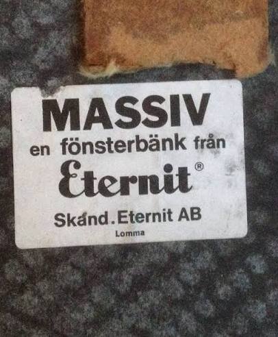 Massiv fönsterbänksetikett från Eternit, Skån. Eternit AB, Lomma, på mörk yta med brunt hörn av material i bildens kant.