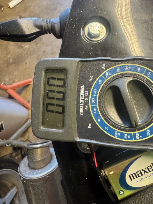 En digital multimeter från Biltema ligger på en svart yta, bredvid ett maxell alkaliskt batteri och en svart kabel.