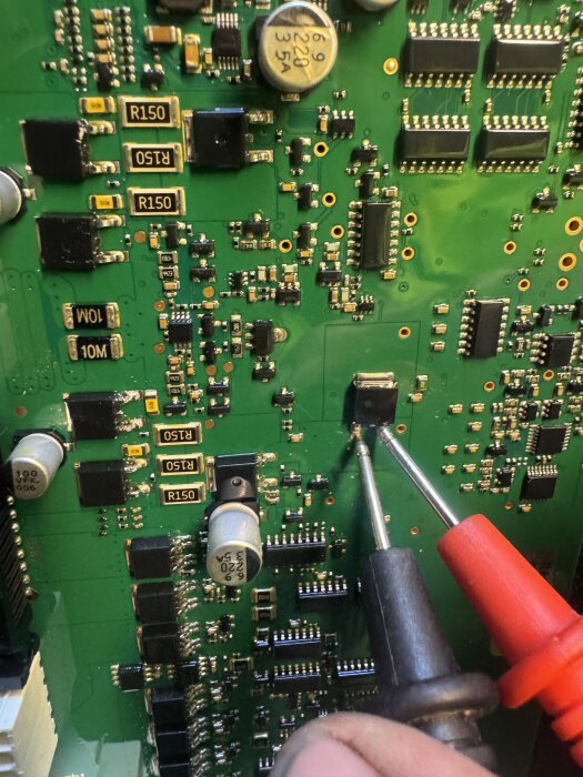 Någon mäter en grön kretskort med multimeterprober, omgiven av olika elektroniska komponenter som motstånd och kondensatorer.