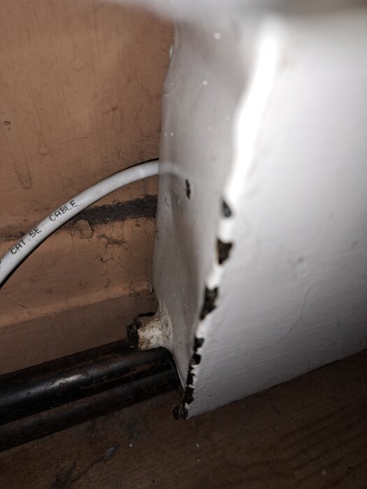 Närbild av ett rör i metall och en sliten vägg med en vit kabel i bakgrunden. Rördelarna är rostiga och målade vita.