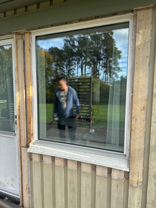 En man reflekteras i ett fönsterglas, omgivningen visar träd och en veranda. Fönstret är del av en husfasad under renovering.