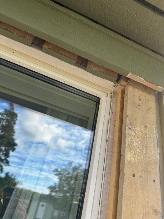 Renovering av fönster med synlig isolering och omålad träpanel på en husfasad under arbete, spegling av himmel och träd i glaset.