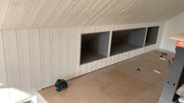 Vit träpanel och inbyggda förvaringsutrymmen under en sned takyta, med några verktyg och byggmaterial på golvet.