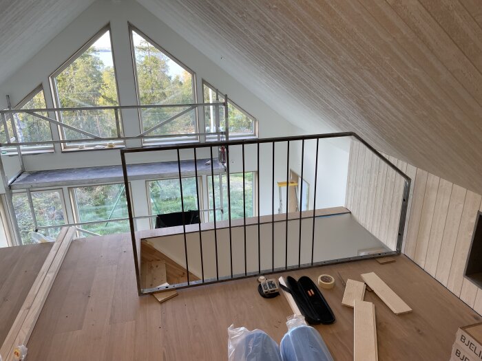 Loftvy med nytt trä- och metallräcke, trägolv och lofttrappa syns. Stora fönster med utsikt över träd utanför, samt byggmaterial på golvet.