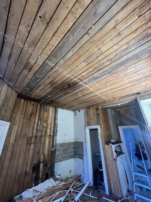 Ett rum med synligt träplanktak och träväggar under renovering, med rivningsmaterial och verktyg på golvet.