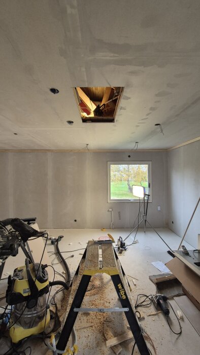 Rum under renovering med hål i taket, verktyg och utrustning utspridda på golvet, och arbetsbänk framför fönster med utsikt mot trädgård.