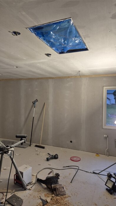 Byggdammigt rum med verktyg och sladdar på golvet. Taket har hål för belysning med blå plastskydd. Väggen saknar färg. Steg i renovering.