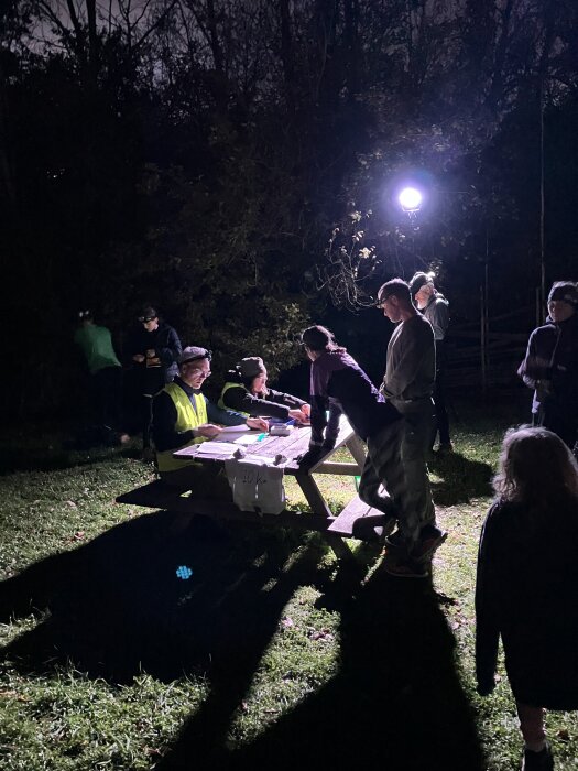 Personer samlade kring ett bord utomhus på natten, belysta av en stark lampa. Några bär reflexvästar och det finns papper och föremål på bordet.