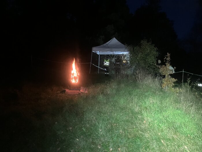 En eld i en eldkorg lyser upp en mörk kväll, med ett vitt partytält och omgivande grönska i bakgrunden.