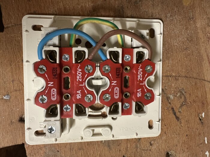 Eluttag med röda terminaler, anslutna med blå, brun och gulgrön kabel. Märkt "16A 250V" och "ELKO". Monterat på trävägg.