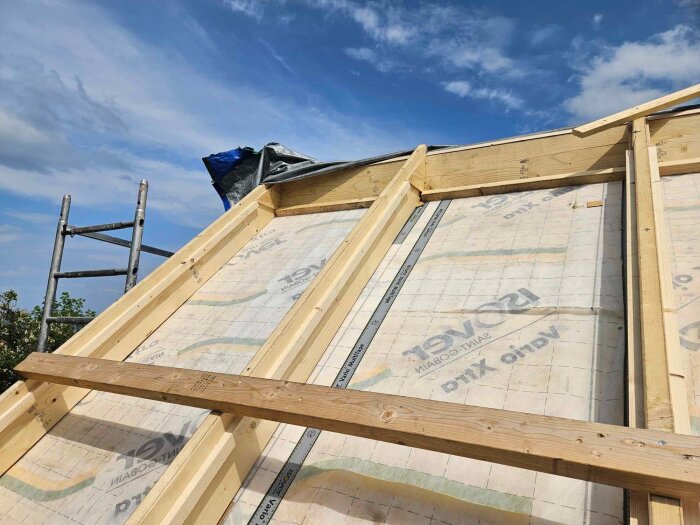 Tak under renovering med synlig ångspärr och träbjälkar, byggställning i bakgrunden, blå himmel ovanför.
