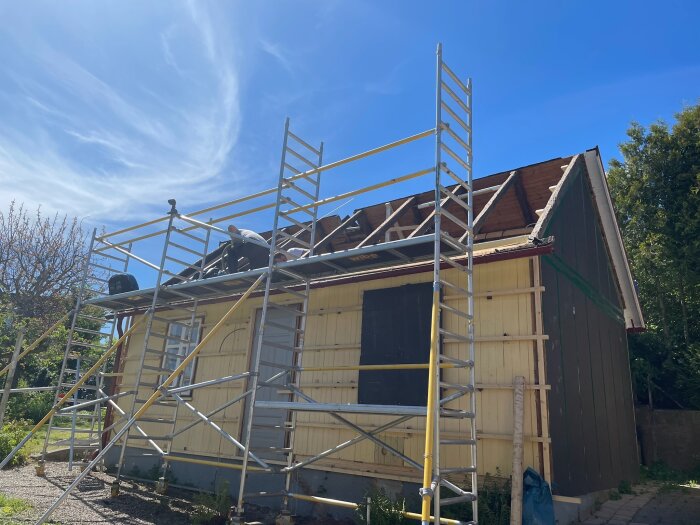 Gult hus under renovering, byggställning monterad längs väggen, takarbeten pågår under klarblå himmel.