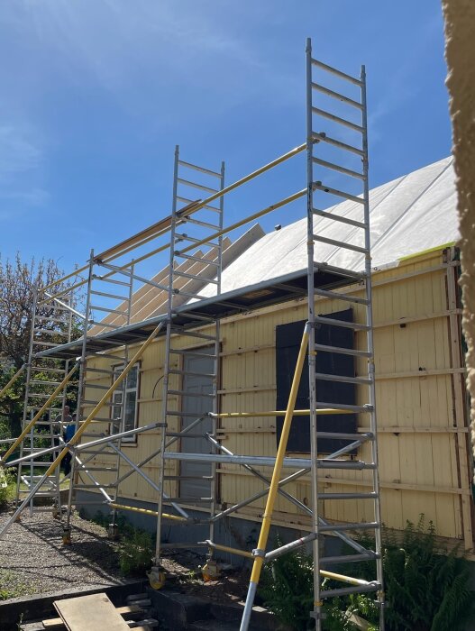 Byggställning vid stuga under renovering, med takarbete pågår. Panel och ångspärr synliga, blå himmel i bakgrunden.