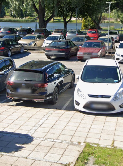 Parkeringsplats med flera bilar, inklusive en svart och en vit bil i förgrunden. Bakom syns träd och en sjö eller damm.