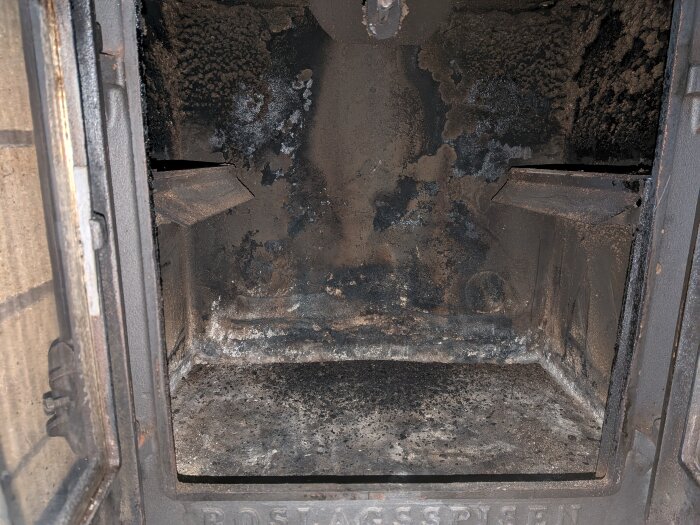 Insidan av en sotig Roslagsspis med öppna dörrar, visar brännkammaren och märkningen "Roslagsspisen" längst ned.