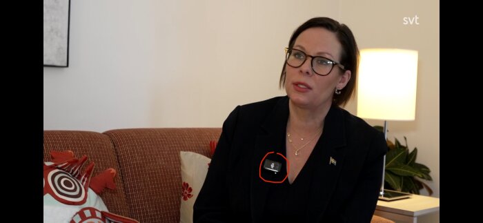 Kvinna med glasögon sitter i soffa, med mikrofon på jackan och svensk flagga synlig.