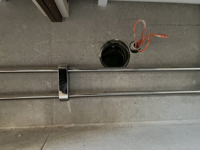 Rund öppning i badrumsvägg, delvis täckt av vattenrör och elsladdar. Problem vid elinstallation med uttag under kommoden.