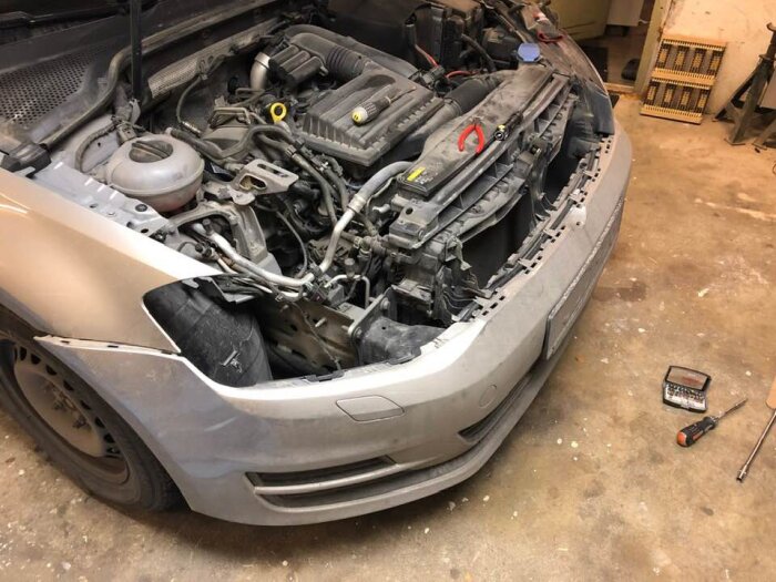 Bil med öppen motorhuv, delar av fronten och lampor borttagna, verktyg ligger på golvet i ett garage.