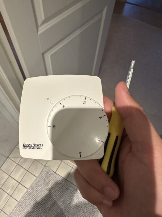 Termostat för vattenburen golvvärme, märkt "Energi Jägarna", med en skruvmejsel i handen, sedd ovanifrån i ett badrum.