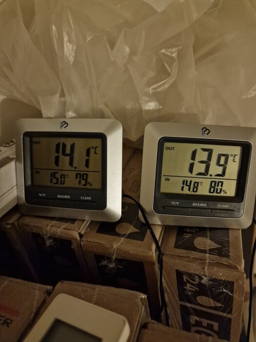Två digitala termometrar visar temperaturer på 14,1°C och 13,9°C med luftfuktighet på 79% respektive 80%, placerade på kartonger med plast i bakgrunden.