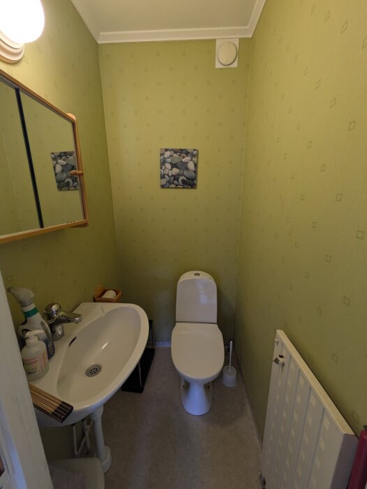 Liten gästtoalett med grön tapet, spegelskåp, handfat, toalettstol, och element. Diskuteras för renovering och förbättring av utrymme och inredning.