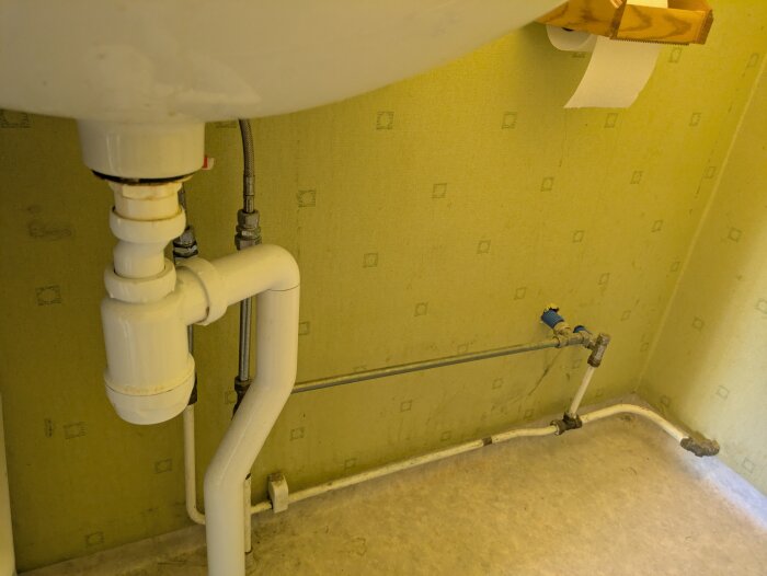 Under ett handfat i en liten gästtoalett, synliga rördragningar mot en gul tapet, med ett golv i ljus plastmatta.