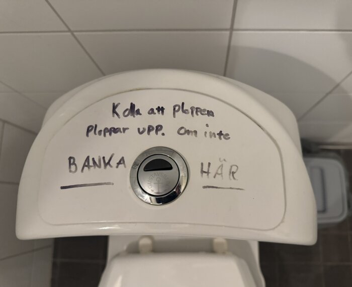 Text på toalettlock lyder: "Kolla att ploppen ploppar upp. Om inte BANKA HÄR", med pil mot knappen.