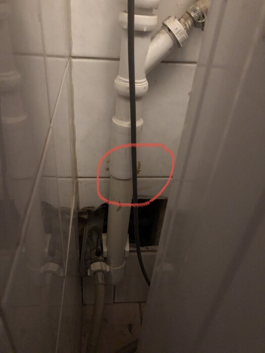 Vitt avloppsrör med rött inringat område som ska bytas ut. Rörförgrening och anslutningar synliga. Tilebeklädd vägg i bakgrunden.