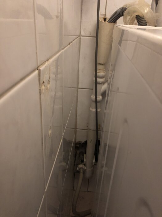 Vit vägg med rörsystem i hörn, synligt från sidan. Ett lodrätt rör och flera anslutningar syns, omgivna av kakelplattor.