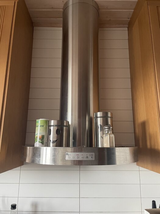 Rostfri köksfläkt omgiven av träskåp, med kryddburkar placerade ovanpå fläkten.