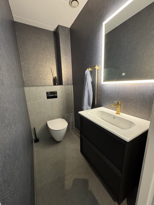 Nyrenoverad modern gästtoalett med mörka väggar, hängande toalett, handfat, guldkran och upplyst spegel.