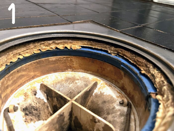 Närbild på duschavlopp med synlig klämring och problem med tätning mot tätskiktet, omgiven av svarta kakelplattor.