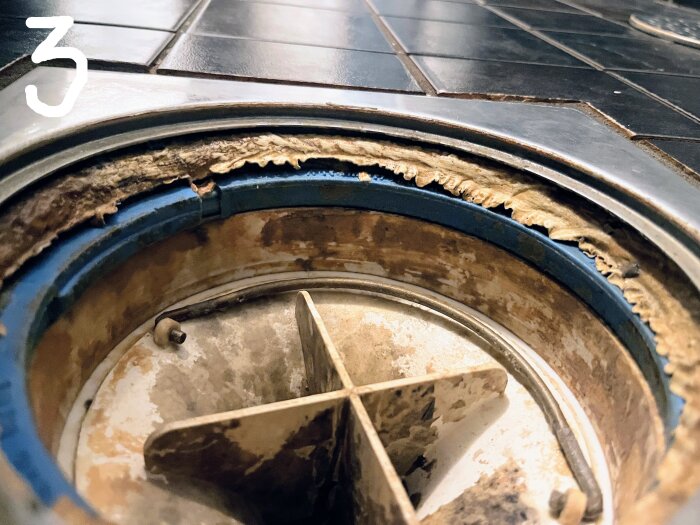 Närbild av duschavlopp med skadad klämring och exponerat tätskikt. Svart kakel på golvet runt avloppet.