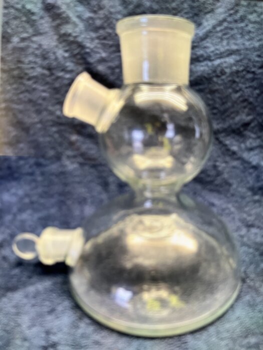 Glasbehållare med dubbla kammare och tre öppningar, används för laboratorieändamål, mot en mörk texturerad bakgrund.